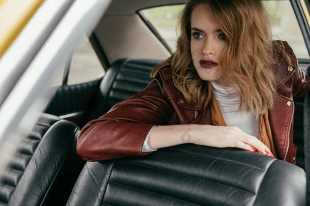 漂亮的年轻女子穿着皮夹克坐在经典的车, 看着远离