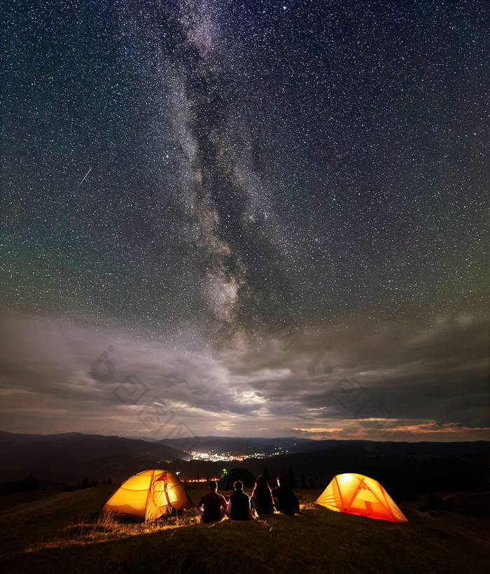 四名徒步旅行者坐在两个橙色帐篷营地, 晚上享受美丽的星空和银河。在远处的城市的灯光坐落在山之间