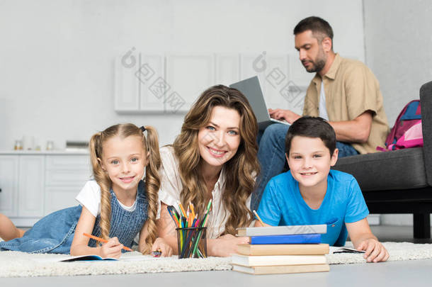 <strong>微笑</strong>的母亲和孩子们看着相机, 同时做家庭作业和人在沙发上使用笔记本电脑在家
