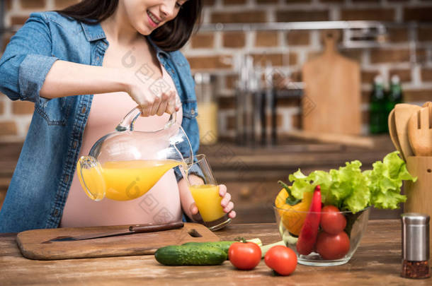 在厨房里把橙汁倒进玻璃杯的微笑怀孕妇女的镜头