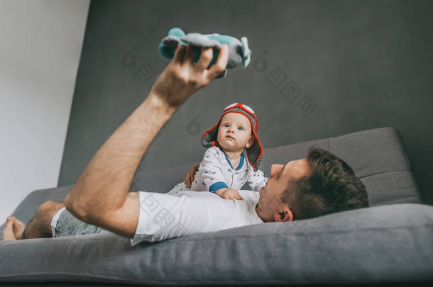 父亲抱着玩具飞机躺在沙发上, 玩着可爱的婴儿戴着针织的飞行员帽