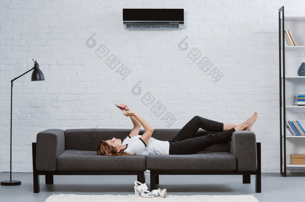 快乐的年轻女子使用智能手机沙发下<strong>空调</strong>挂在墙上