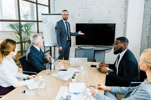  business coach in formal wear gesturing near tv with blank screen and multicultural coworkers 