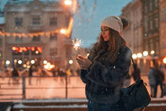 美丽的年轻妇女在毛皮大衣持有烟火享受冬季圣诞节心情在老雪欧洲城市与室外溜冰场背景