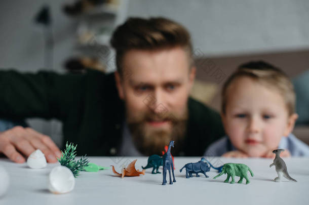 选择焦点的父亲和可爱的小儿子看着被安排的玩具恐龙在桌面上一起在家