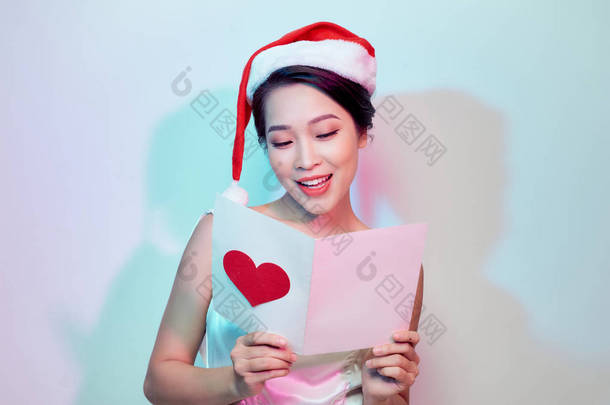 美丽的年轻女子在圣诞老人帽子拿着明信片与红色心脏在光背景