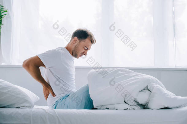 年轻人穿着<strong>睡衣</strong>坐在床上, 背痛痛苦的侧面观察