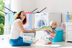 母亲和孩子在洗衣房里用洗衣机或滚筒烘干机。家庭琐事。现代家庭设备和洗涤洗涤剂在白色阳光的家。烘干机架清洗洗衣服. 