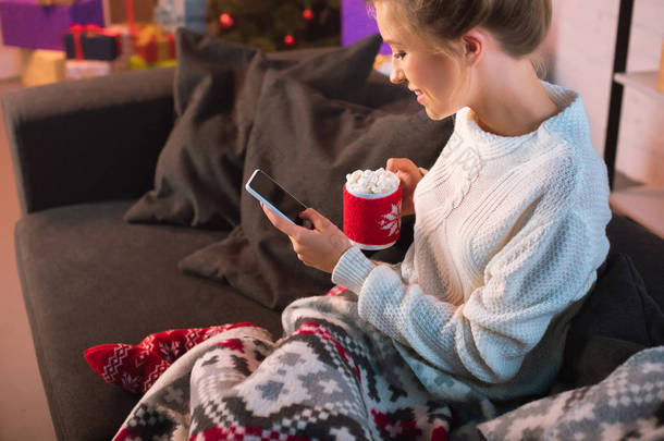 年轻的金发碧眼的女人拿着一杯热可可与棉花糖和使用智能手机在圣诞节时间