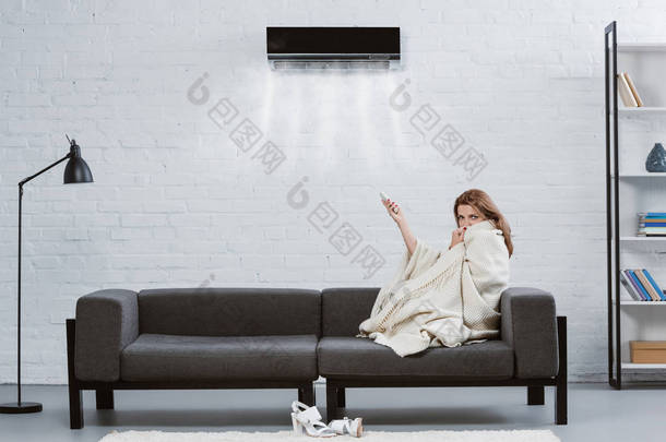 裹着毯子的年轻妇女在沙发<strong>下</strong>挂在墙上的空调和吹冷的空气
