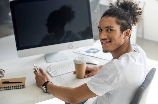 年轻的非洲裔美国人与<strong>纸杯</strong>和智能手机微笑着在工作场所的相机