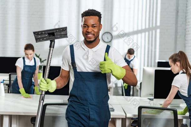微笑着的非洲裔美国清洁工拿着真空吸尘器刷子，在同事们身边展示大拇指