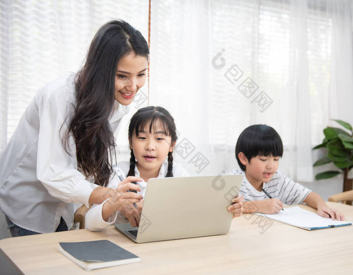 在家里客厅里，一位年轻的亚洲女人坐在桌子旁边，用笔记本电脑帮她儿子和女儿做作业.