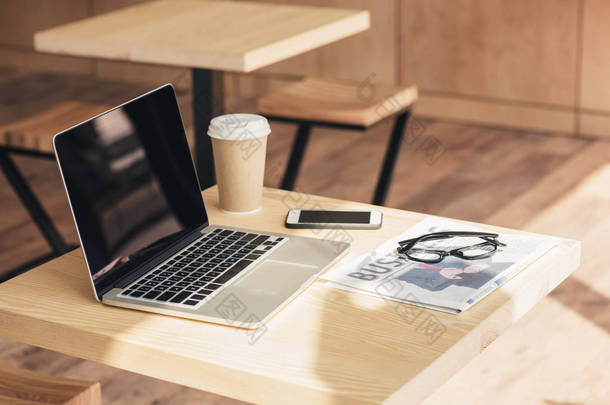 带空白屏幕的笔记本电脑、智能手机和商务报纸在咖啡店的餐桌上