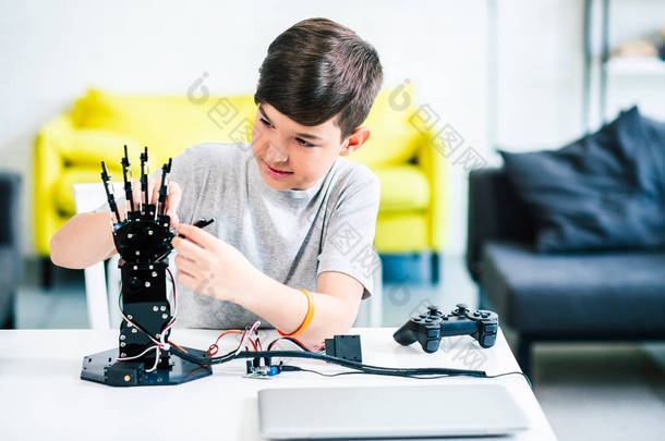 热情的聪明男孩坐在桌旁，同时尝试机器人手和准备<strong>工程</strong>课程