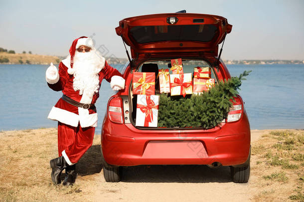 正宗的圣诞老人靠近汽车与开放的树干充满了礼物和杉树户外