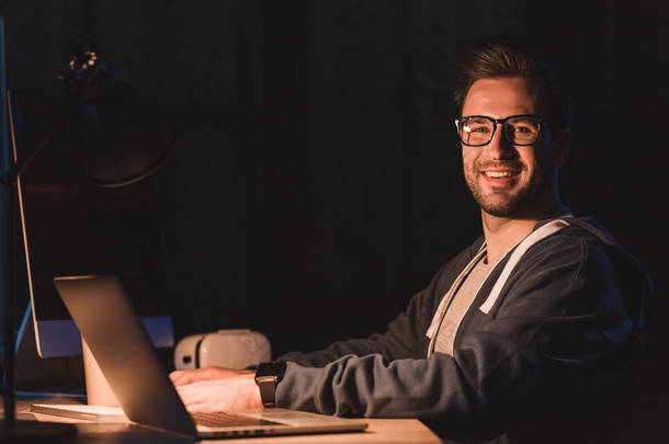 英俊年轻的程序员在眼镜微笑, 而在夜间使用笔记本电脑和台式电脑的相机