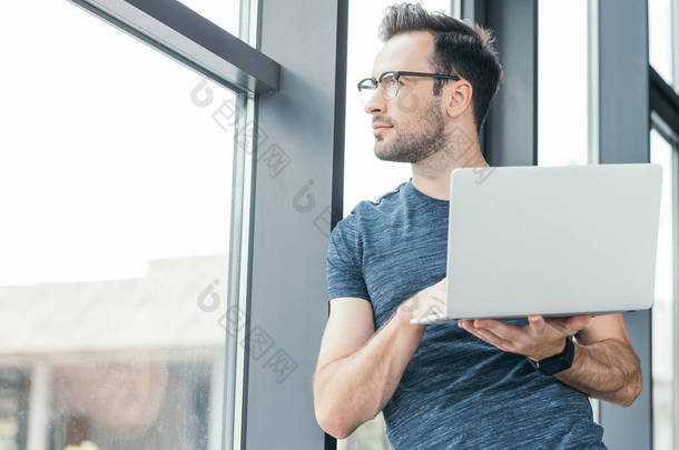 英俊的年轻男子在眼镜持有笔记本电脑和看窗口