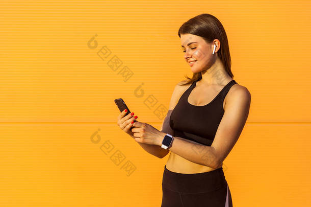 年轻的快乐有吸引力的运动女人在黑色运动服使用智能手机,而站在橙色墙背景在阳光下 