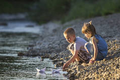 两个可爱的金发孩子, 男孩和女孩在河岸上送水白纸船在明亮的夏天模糊的蓝色背景。快乐童年和户外活动的乐趣和游戏概念.