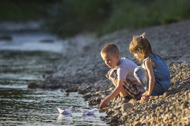 两个可爱的金发孩子, 男孩和女孩在河岸上送水白纸船在明亮的夏天模糊的蓝色背景。快乐童年和户外活动的乐趣和<strong>游戏</strong>概念.