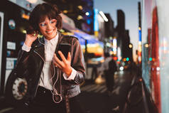 开朗漂亮的年轻女子在凉爽的眼镜和时髦的穿着步行在大都会街与夜灯享受歌曲从播放列表在耳机和阅读 sms 与好消息智能手机