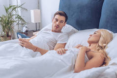 可疑的男人躺在床上与智能手机和看着睡觉的女朋友, 不信任的概念 