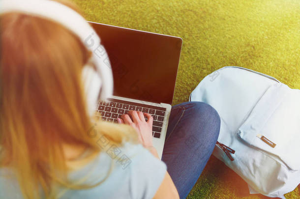 高视角<strong>的</strong>年轻女子坐在<strong>草</strong>地上耳机头上和使用笔记本电脑