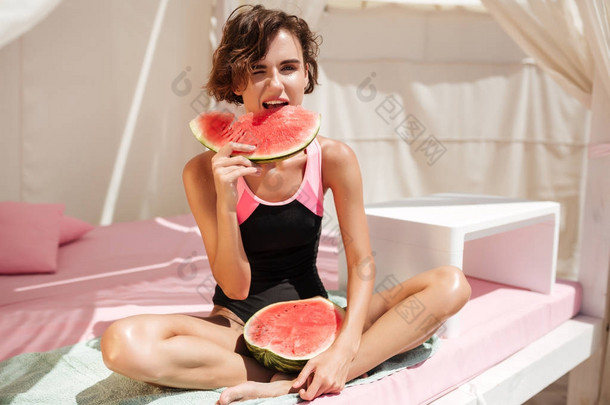 美丽的女孩在时髦的泳装坐在沙滩帐篷, 并期待在相机与<strong>西瓜</strong>切片手。花时间在海滩上<strong>吃西瓜</strong>的年轻女士画像