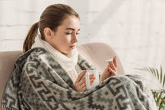 生病的女孩，裹在毯子里，看着温度计，端着一杯热饮