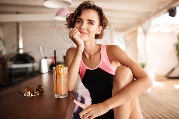 年轻漂亮的女孩在泳衣的肖像坐在海滩酒吧与太阳镜和鸡尾酒<strong>柜台</strong>。美丽的微笑的女士坐在酒吧<strong>柜台</strong>与鸡尾酒和朦胧的摄像头看