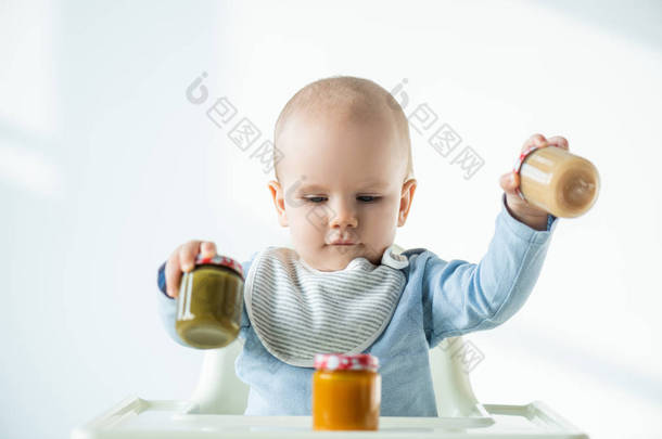 Selective focus of baby holding jars of vegetable baby nutrition while sitting on feeding chair on w