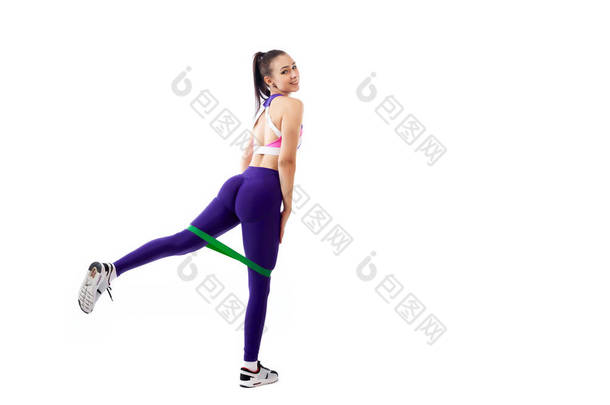 一个在运动紫色短上衣和健身房紧身裤的伊蓬女子教练在脚前冲刺与运动健身橡皮筋, 舒展腿在白色隔离的背景在演播室 