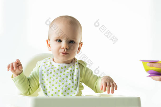 Adorable baby looking at camera while sitting on feeding chair near mother with bowl of baby nutriti