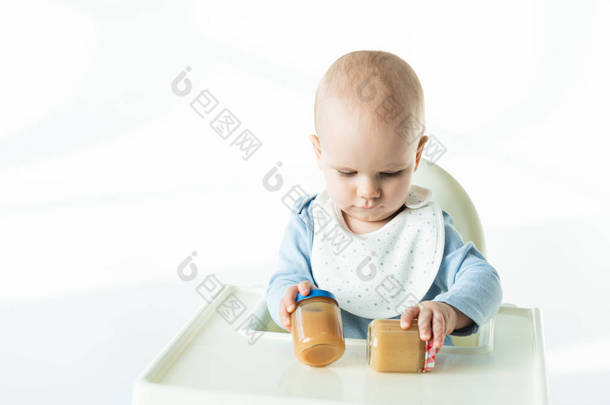 Adorable baby boy holding jars with baby food on table of feeding chair on white background