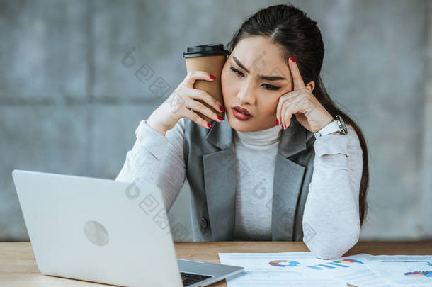 疲惫的年轻女商人拿着咖啡去, 并在工作场所使用笔记本电脑