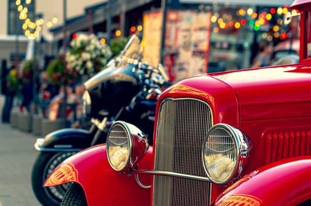 在现代摩托车的背景下，一辆带有彩色前灯的老式红色轿车出现在夏季街道上。拉脱维亚里加