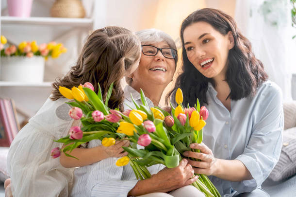 祝你在妇女节快乐!小女儿向妈妈和奶奶表示祝贺, 给她们送花郁金香。奶奶, 妈妈和女孩微笑着拥抱。家庭假期和团聚.