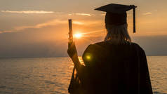 在海边的夕阳背景上，一位拿着文凭的大学毕业生的后视图。高等教育的新机遇