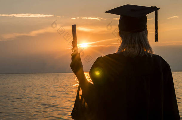 在海边的夕阳背景上，一位拿着文凭的大学毕业生的后视图。高等教育的新机遇