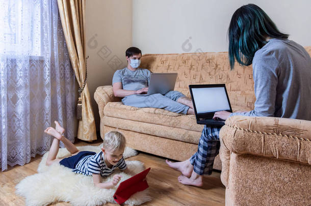 一个年轻的家庭独自呆在家里。戴着面具的男人坐在沙发上，在笔记本电脑上工作。女人