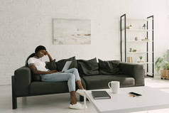 体贴的非裔美国自由职业者坐在沙发上，在宽敞的客厅里拿着笔记本电脑