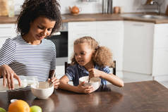 与女儿共进早餐时，一位身穿条纹t恤的年轻非裔美国妇女正在往碗里倒牛奶