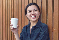 微笑的年轻女士站在户外的木墙上，手里拿着一杯咖啡。