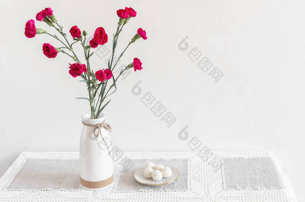 陶瓷花瓶里的桌子上有红色的<strong>康乃馨</strong>，旁边是一盘糖果。白色背景斯堪的纳维亚风格前视图，复制空间.