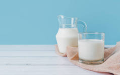 天然鲜牛奶在玻璃杯和水壶里，放在餐巾上，放在蓝色背景的木制桌子上。饮食促进健康