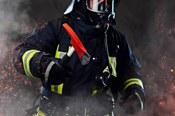 一名身穿制服和氧气面罩的消防员手持一把红色斧头站在火的火花和烟雾在黑暗的背景.