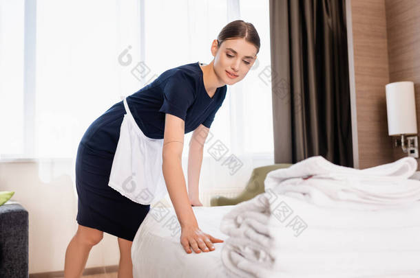 女服务员穿着<strong>围裙</strong>在旅馆房间的毛巾旁边铺床 