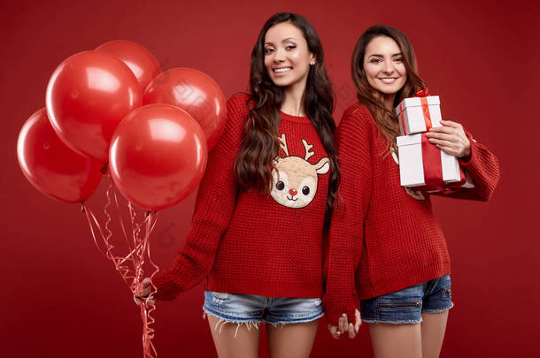 两个穿着时尚舒适冬季毛衣，头戴派对气球，头戴红色背景礼品盒的<strong>非常</strong>疯狂的好朋友双胞胎的画像