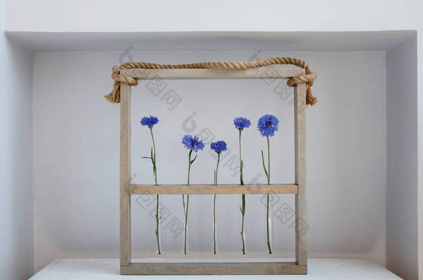 五朵蓝色的漂亮的玉米花，装在一个木制框架的小花瓶里，上面挂着<strong>灰色</strong>的、简单的<strong>花朵</strong>装饰